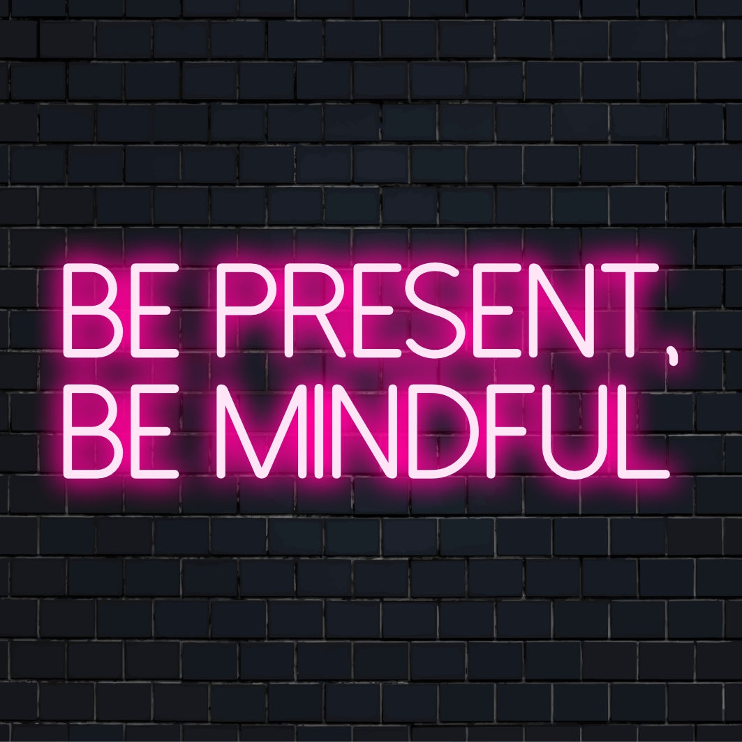 Auf dem maßgefertigten LED-Neonschild steht in leuchtenden, lebendigen Buchstaben „Be Present, Be Mindful“; personalisierte Neon-Wandkunst.