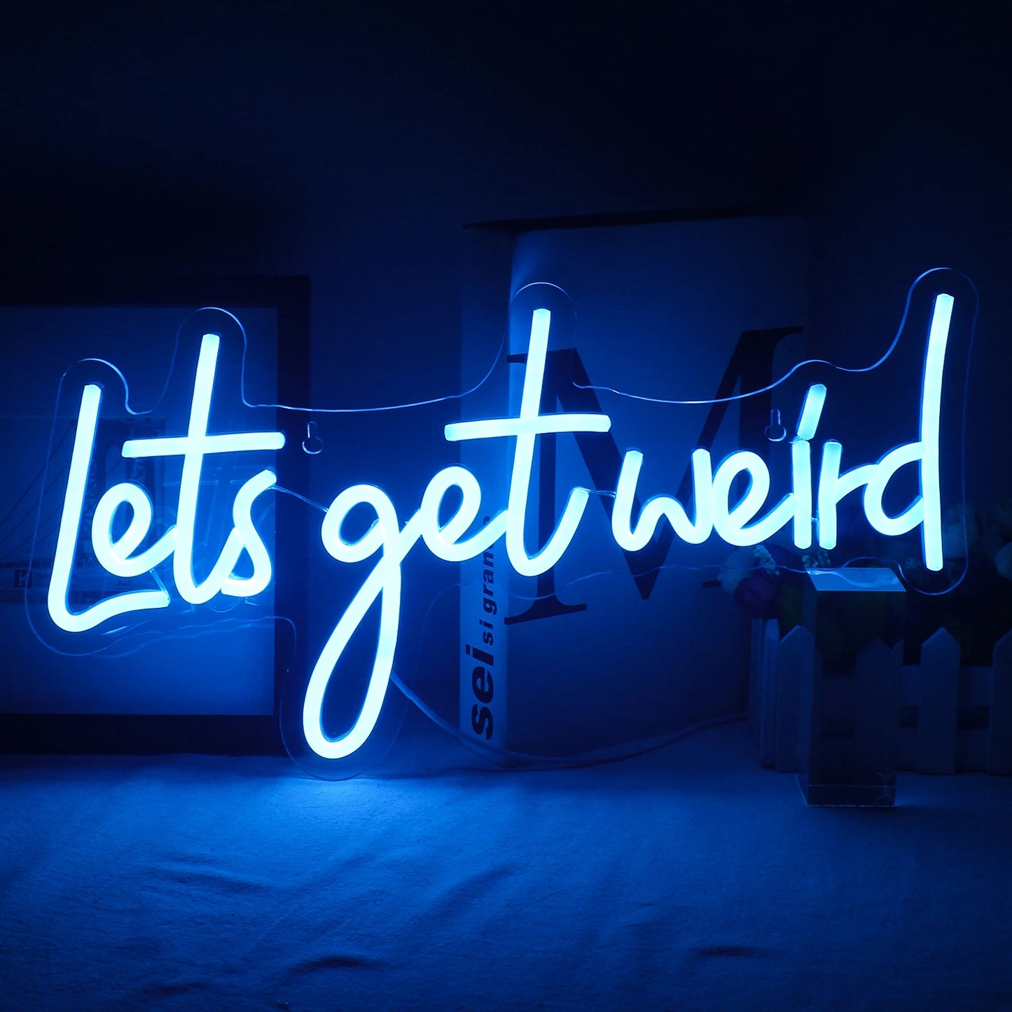 Das Bild zeigt ein leuchtendes „Let’s Get Weird“-LED-Neonschild, das an einer Wand montiert ist.