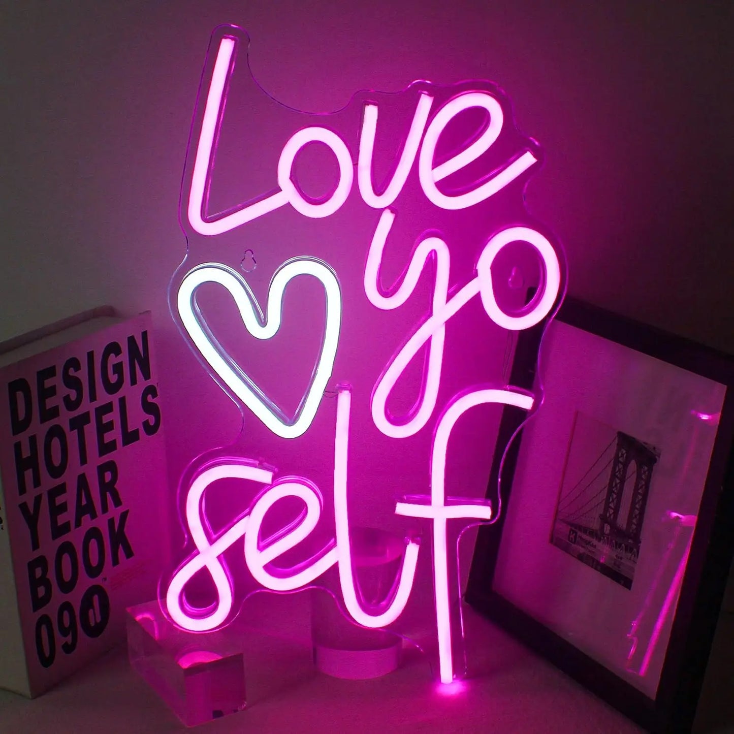 Vor einem einfachen, dunklen Hintergrund wird ein leuchtendes LED-Neonschild mit der Aufschrift „Love Yo Self“ angezeigt.