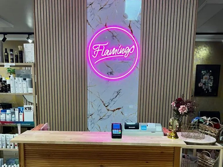 A retail storefront counter with a wooden reception desk, a pink neon sign reading 'Flamengo' on a marble panel, and shelves of products in the background.