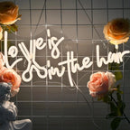 The image features a vibrant, handmade LED neon sign spelling out the phrase "Love is in the Hair.