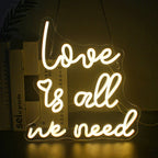 The image features a vibrant LED neon sign that reads "Love is all we need" in a flowing, cursive font.