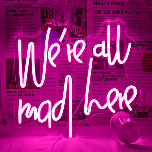 A glowing LED neon sign displaying the phrase "We're All Mad Here" in a whimsical, cursive font.