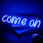 The image features a vibrant LED neon sign displaying the words "Come On" in a bold and playful script font.