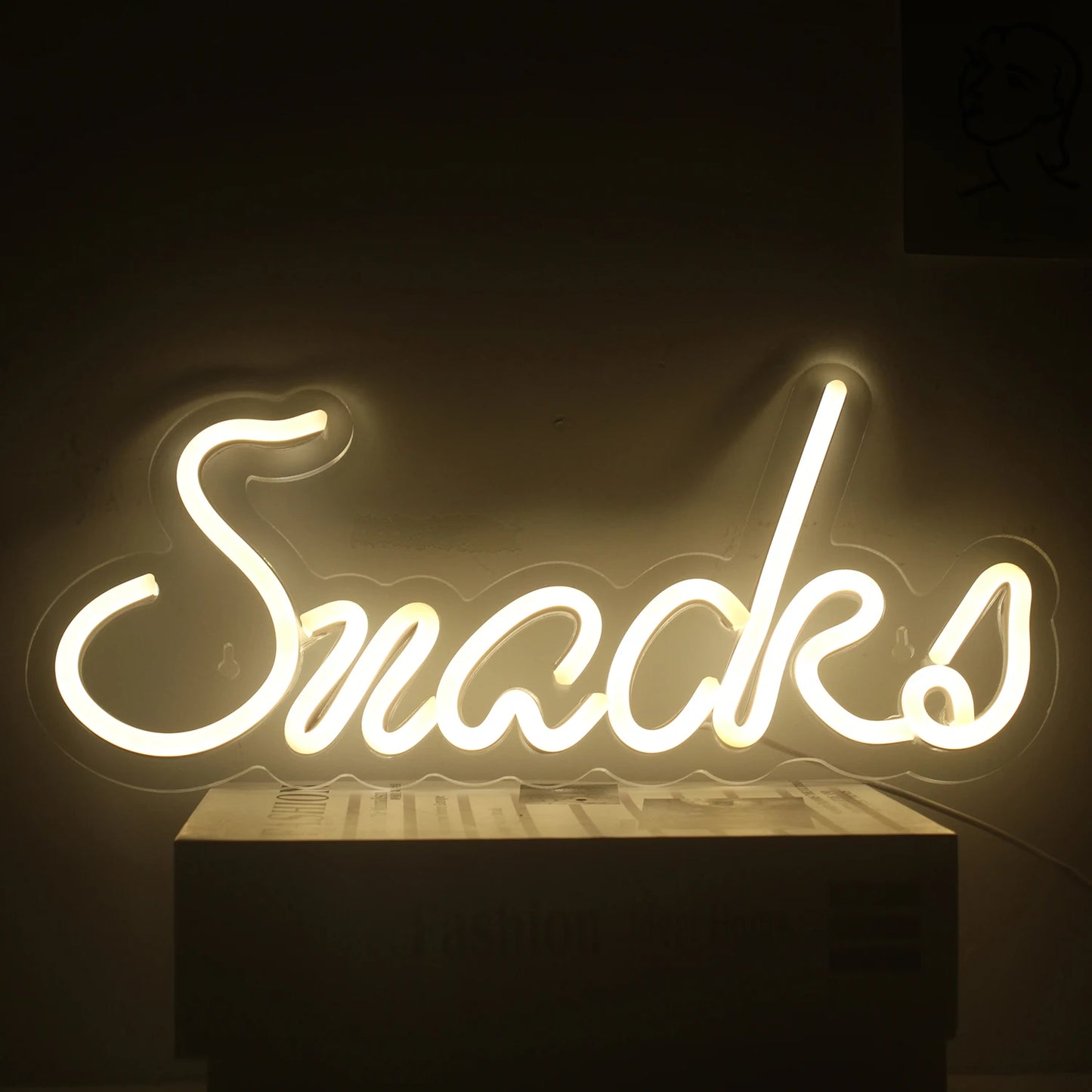 Das Bild zeigt ein leuchtendes LED-Neonschild in Form des Wortes „Snacks“ in einer verspielten, kursiven Schriftart.
