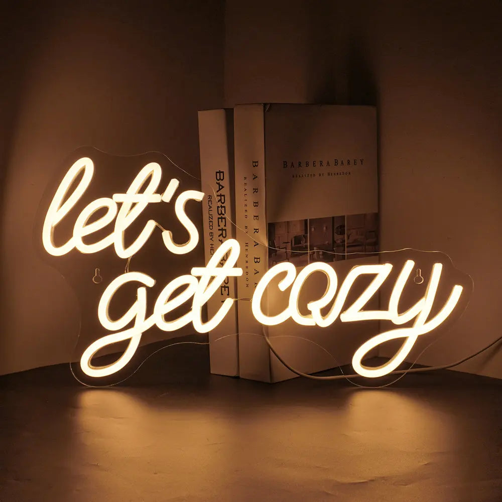 Das Bild zeigt ein leuchtendes LED-Neonschild mit dem Satz „Let’s Get Cozy“ in einer warmen, einladenden Schreibschrift.