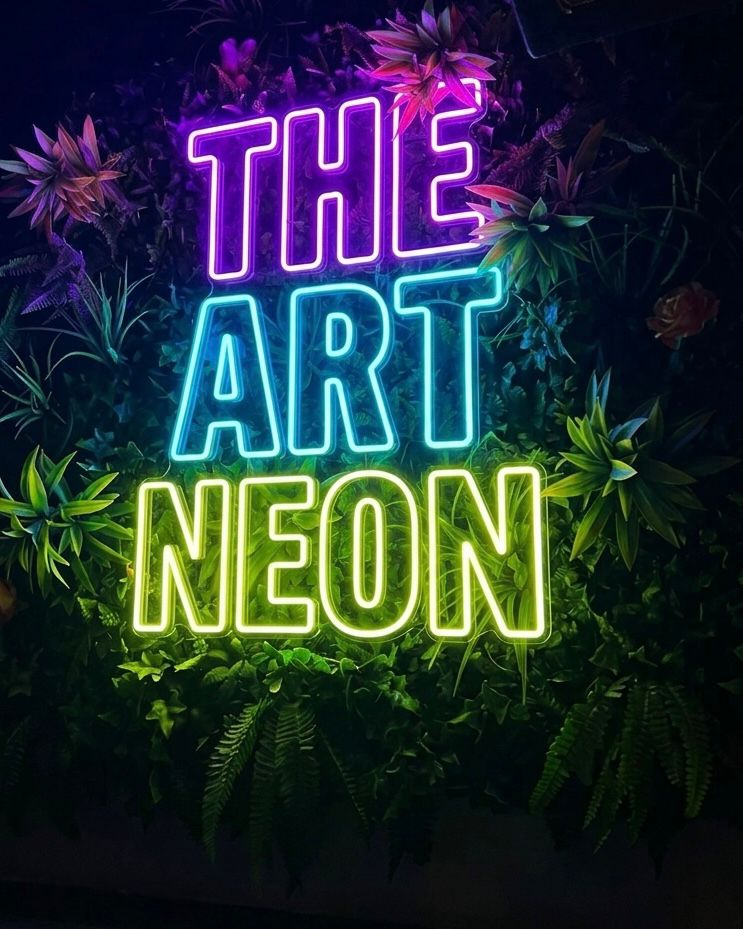 Mehrfarbiges LED-Neonschild von The Art Neon zwischen Pflanzen
