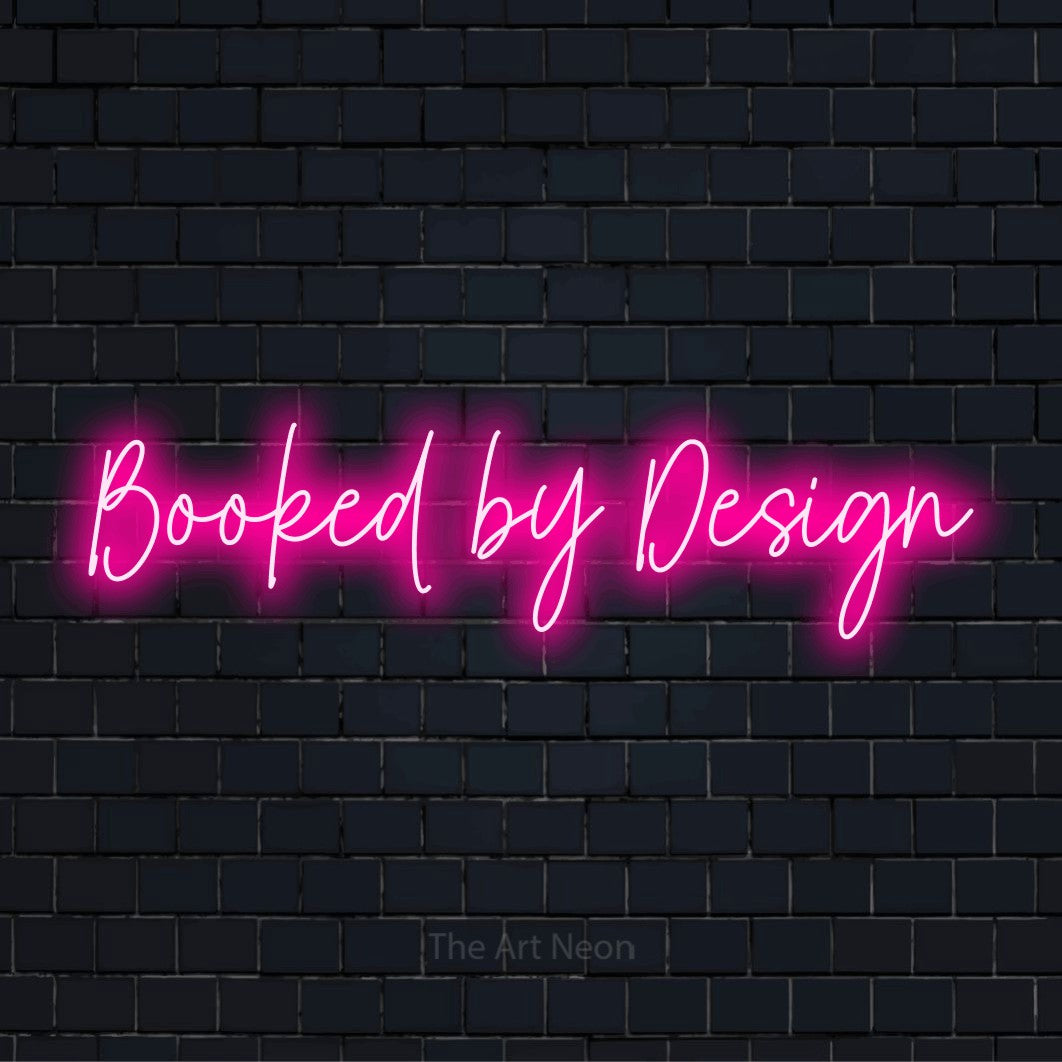 "Booked By Design" script-style LED neon sign on a black brick wall