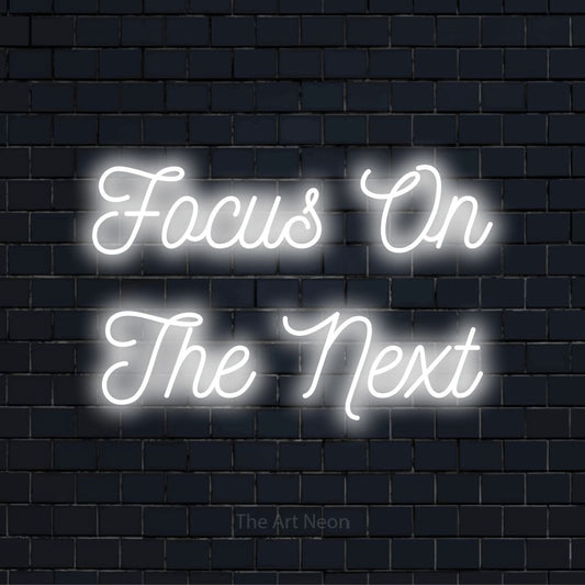 "Focus On The Next" LED neon sign on a black brick wall