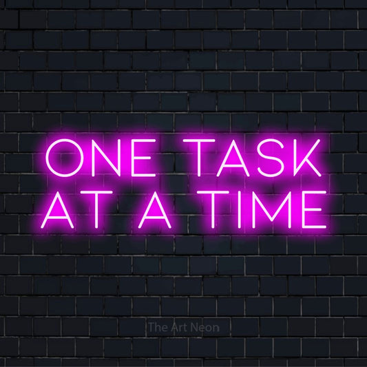 "One Task At A Time" neon text design on a black brick wall