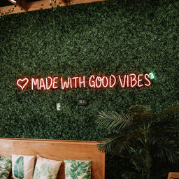 Neon sign 'MADE WITH GOOD VIBES' on a green wall with wooden bench and plants.