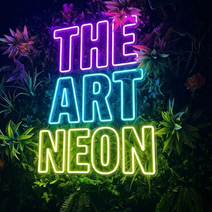 A glowing neon sign reading 'THE ART NEON' in purple, blue, and green lettering, mounted against a dark wall covered with artificial plants and flowers.